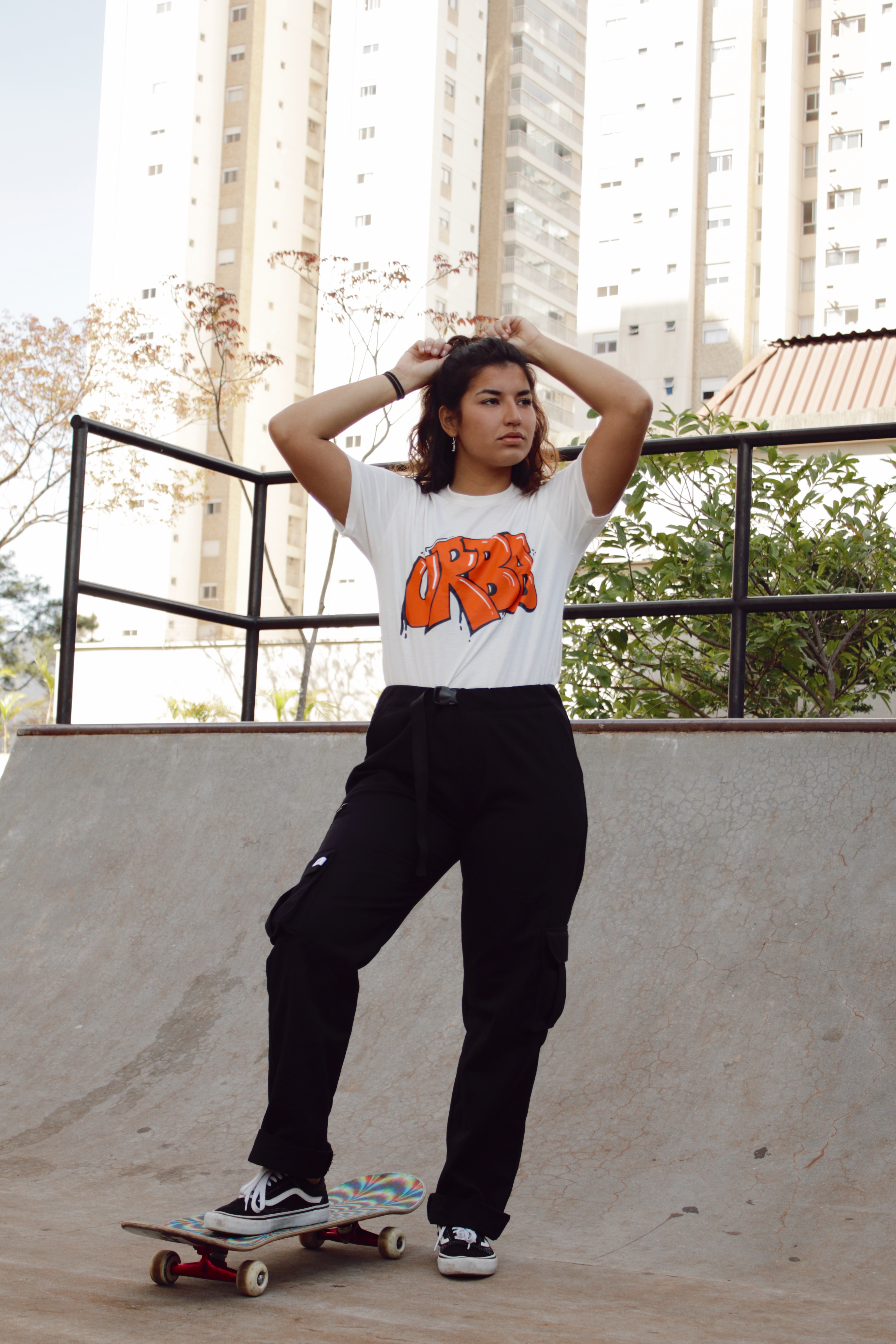 Thais Gazarra em mini rampa de skate usando camiseta branca, calça cargo preta, com um dos pés apoiado no skate e mexendo no cabelo com as duas mãos para trás