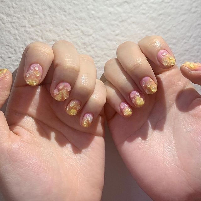 Foto de duas mãos com o esmalte degradê de rosa e amarelo com detalhe de gota de água em gel 3D.