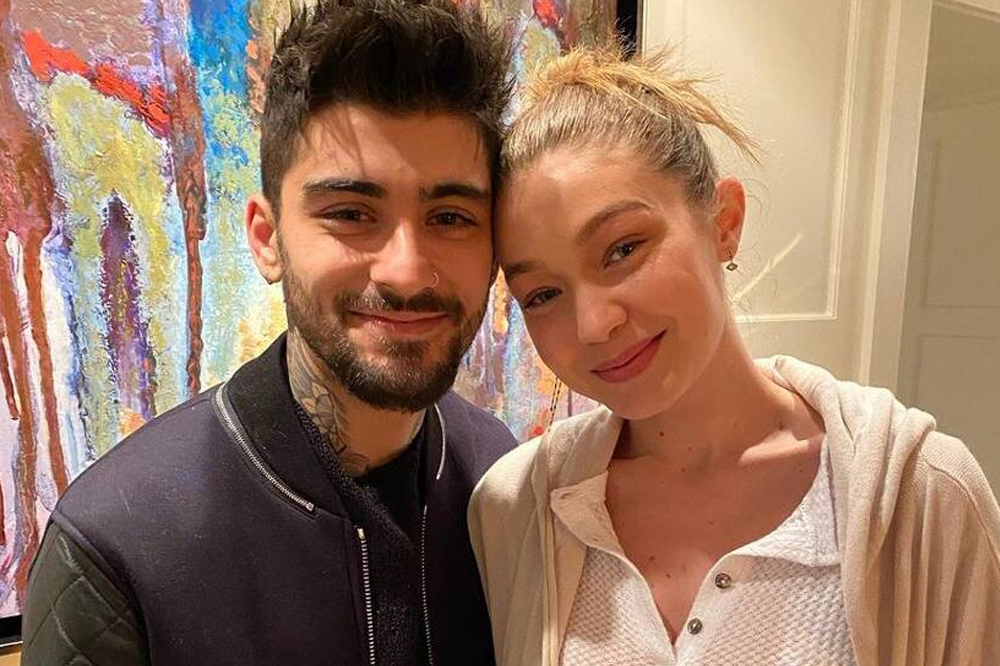 Zayn e Gigi Hadid sorrindo levemente para foto; ele usa uma blusa preta com jaqueta também preta enquanto a modelo está com o cabelo preso e usa uma camisa branca com botões próximos da gola e um casaco bege; eles estão olhando para a câmera com os rostos encostados; ao fundo é possível ver um quadro colorido e uma porta branca atrás de Gigi