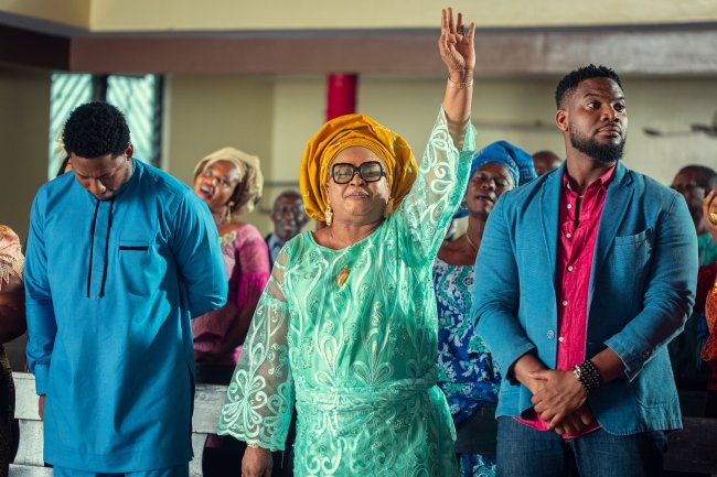 A Naija Christmas Uma mulher com roupas coloridas e a mão levantada no meio de muitas pessoas com roupa azul.