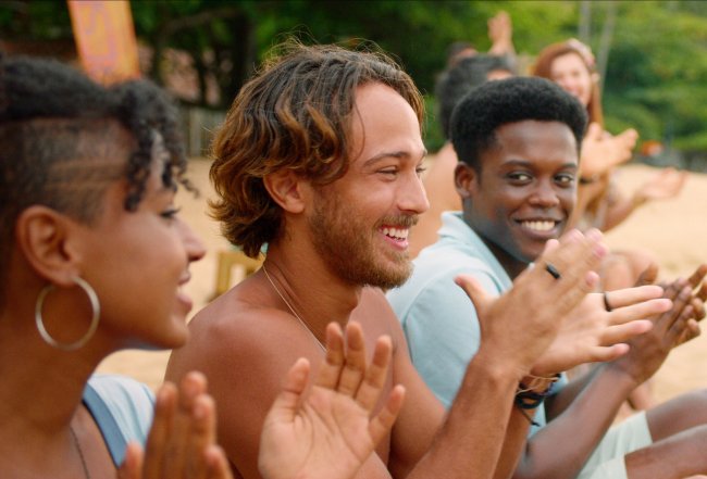 TDV_103_00_40_31_04R Elenco de Temporada de Verão, a nova série da Netflix; os personagens estão sentados na praia alinhados batendo palmas e sorrindo; a foto foi dirada de perfil e é possível ver areia e folhas de árvores verdes ao fundo