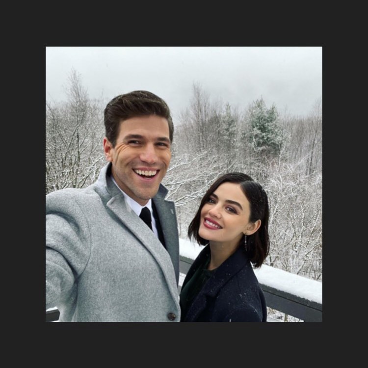 Austin Stowell e Lucy Hale nos bastidores das gravações de O Jogo de Amor/Ódio; eles estão tirando uma selfie sorrindo e Austin segura a câmera; a paisagem ao fundo são montanhas e árvores cobertas por neve; Austin usa um casaco cinza com terno e gravata por baixo e Lucy usa um casaco preto; a borda da imagem é um quadrado preto