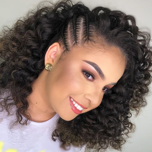 cabelo-cacheado-com-tranca Selfie de uma mulher de perfil. Ela usa uma camiseta cinza, maquiagem com sombra rosa, brinco dourado e cabelo cacheado solto com tranças laterais. Ela está de perfil, olha para frente e sorri.