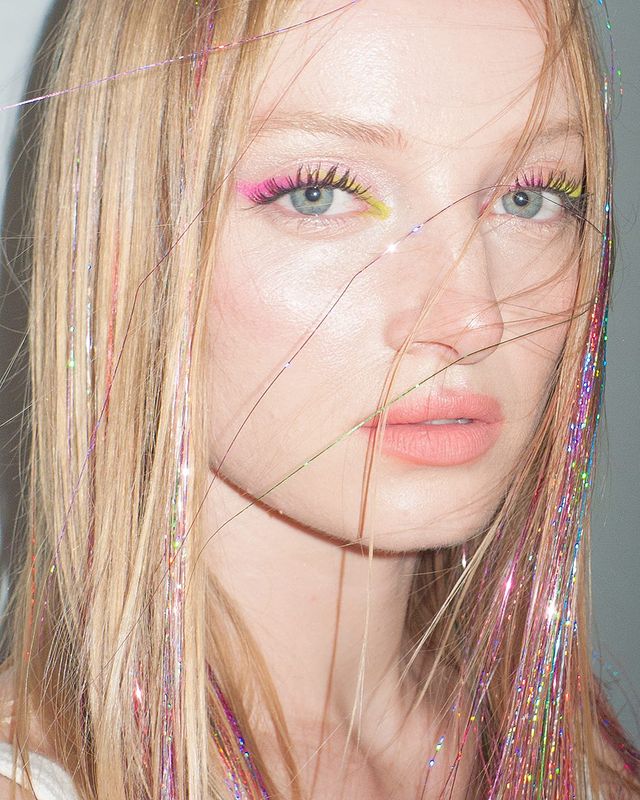 cabelo-com-fios-de-glitter Foto de uma mulher. Ela está com uma maquiagem com sombra rosa e cabelo solto com fios de glitter coloridos. Ela olha para a câmera e não sorri.