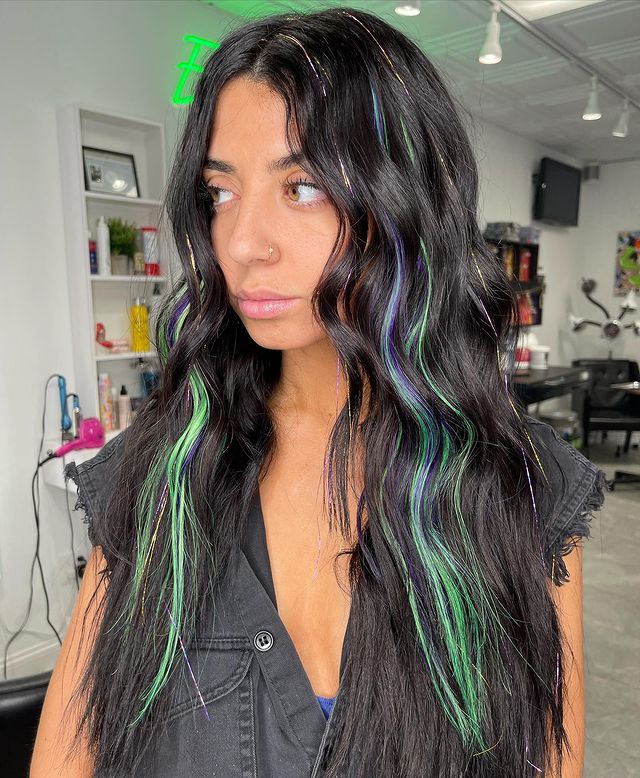 cabelo com fios de glitter Foto de uma mulher em um salão de beleza. Ela usa uma camisa preta e cabelo preto solto e ondulado com pequenas mechas com fios de glitter verdes. Ela olha para o lado e não sorri.