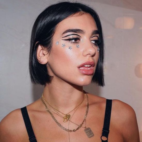 Foto da cantora Dua Lipa. Ela usa um top preto de alcinha, colares, cabelo solto e maquiagem com delineado preto no formato banana. Ela olha para o lado e não sorri.