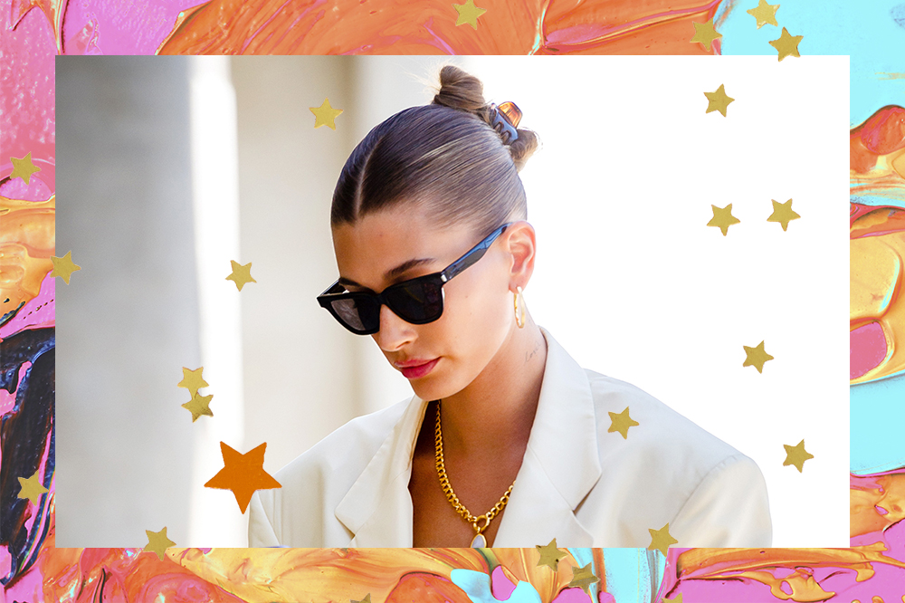 Hailey Bieber olhando para baixo usando óculos de sol e com cabelo preso com piranha em coque. O fundo da montagem possui tintas laranjas, rosas e azuis, além de estrelinhas douradas