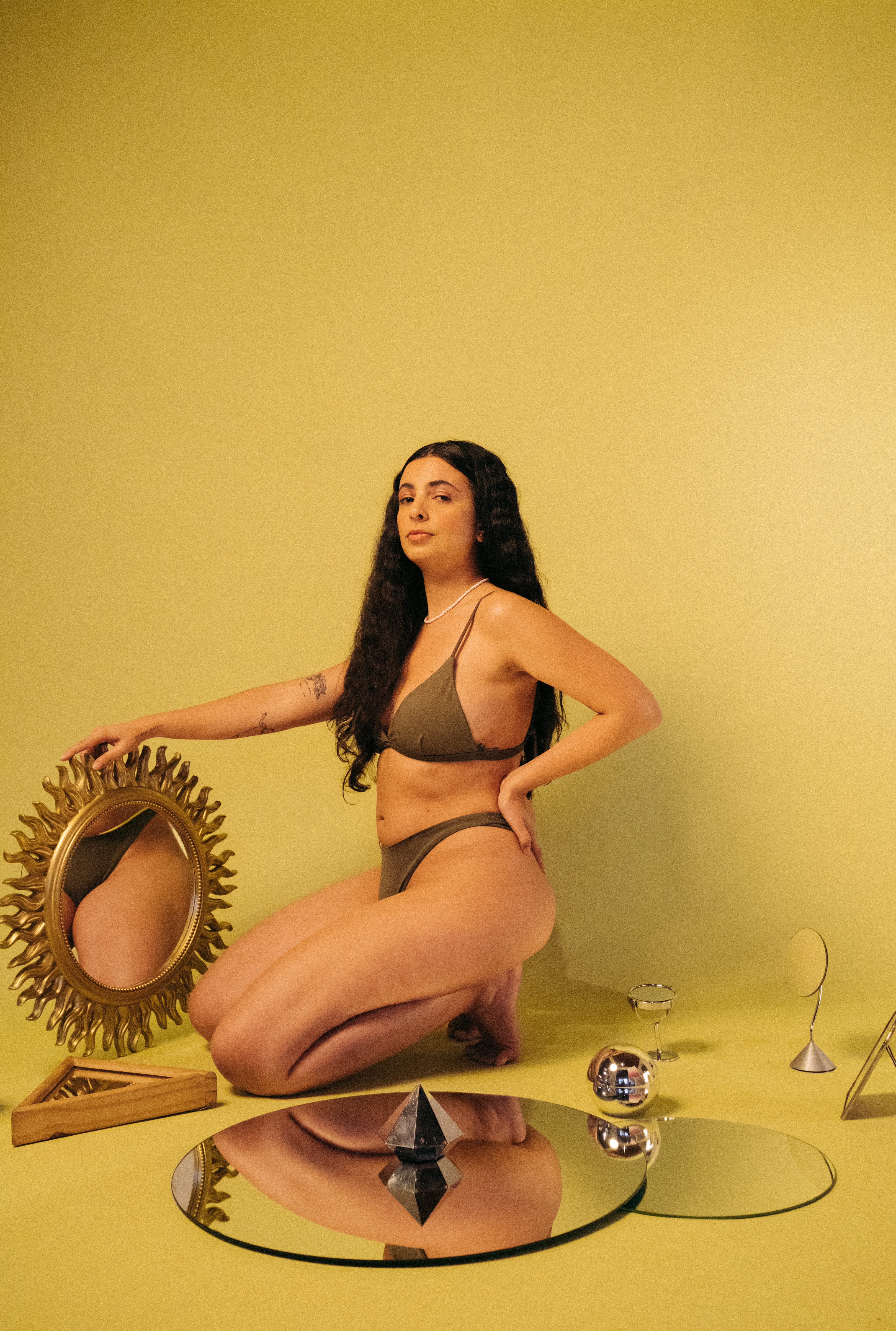 lela-brandao-biquinis-colecao-espelhos Ilustradora Lela Brandão usando biquíni marrom da sua própria marca, agachada com uma das mãos no quadril, segurando um espelho e com expressão facial séria