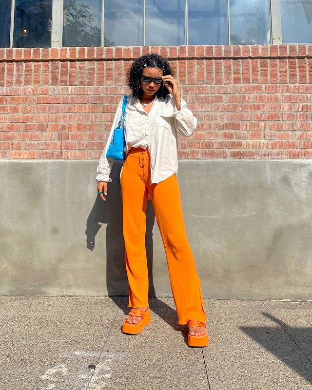 look-com-camisa-branca-e-calca-colorida Foto de uma mulher em uma calçada. Ela usa uma camisa branca de manga longa, calça laranja, tamanco laranja e shoulder bag azul. Ela está com o cabelo cacheado e curto solto, de óculos de sol, olha para o chão e não sorri.
