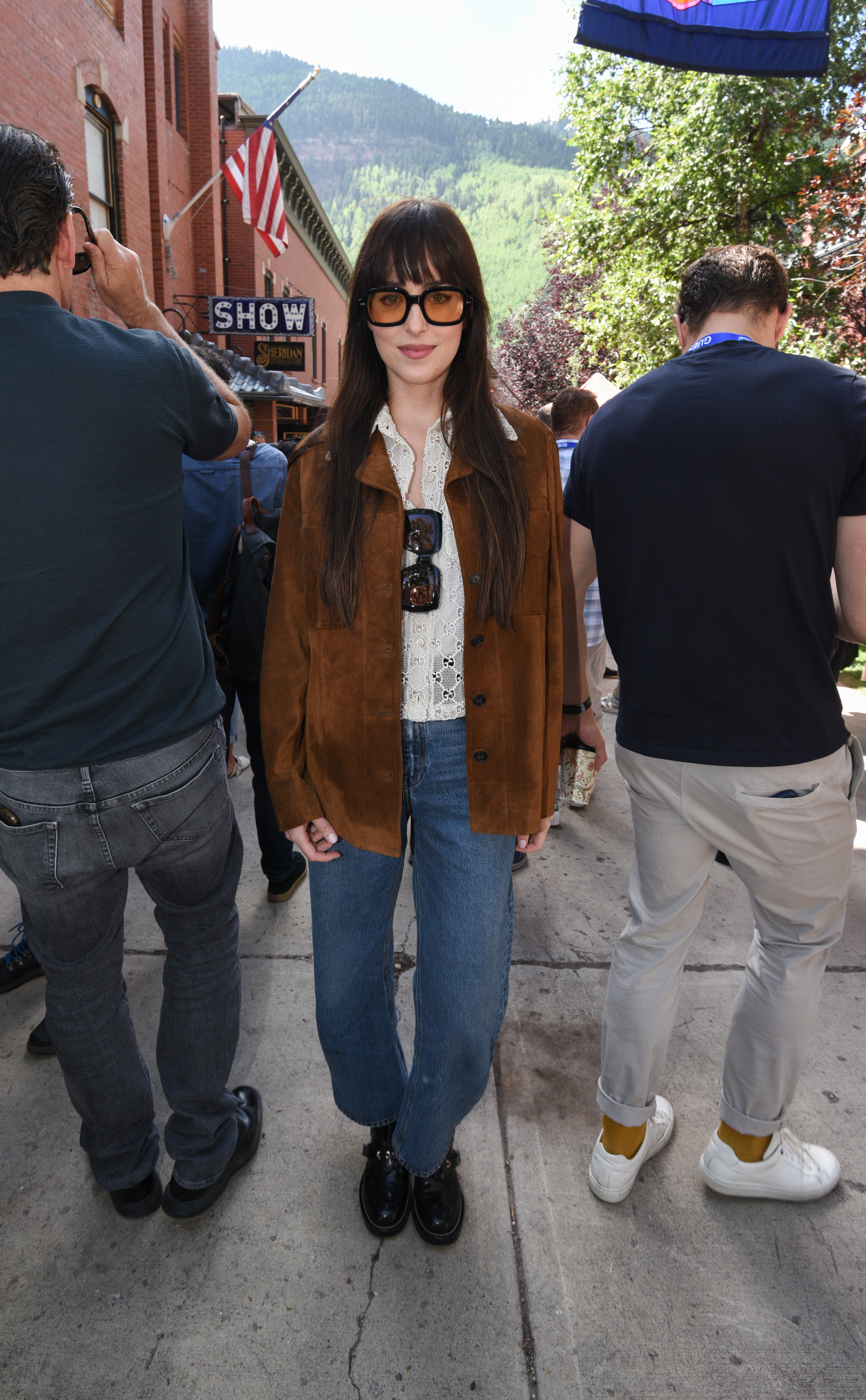 2021 Telluride Film Festival Dakota Johnson usando óculos de lentes amareladas, camisa branca, jaqueta marrom de camurça, calça jeans e bota preta na rua, ao lado de pessoas viradas de costas