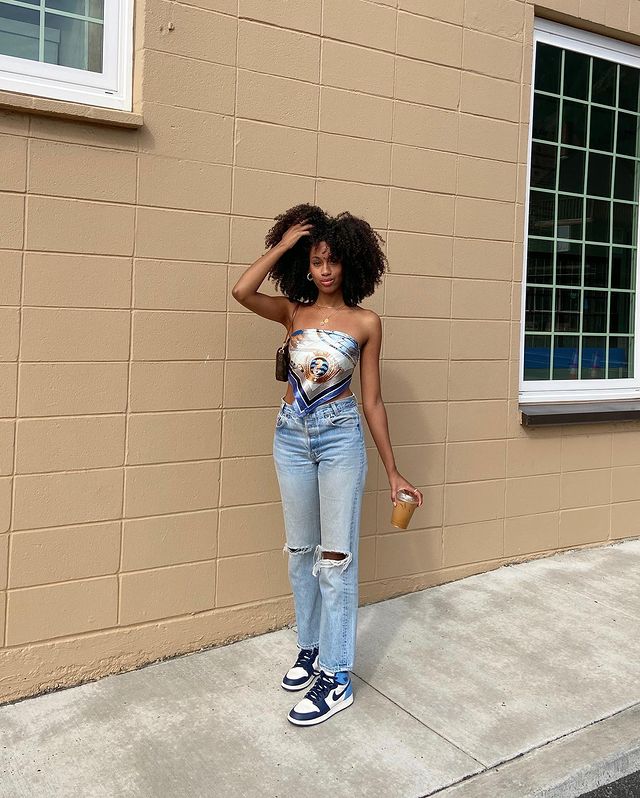 Foto de uma mulher em uma calçada. Ela usa um top feito com um lenço, uma calça jeans, tênis com detalhes em tom de azul e shoulder bag. Ela segura um copo com a mão esquerda e está com a mão direita no cabelo, olha para a câmera e não sorri.