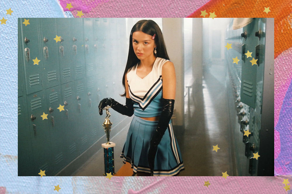 Foto de Olivia Rodrigo, com roupa de líder de torcida azul com branco, presilhas no cabelo, luvas pretas longas de couro e um troféu.