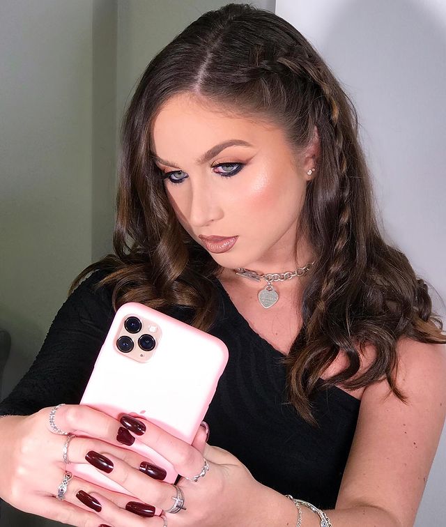 penteado-cabelo-tranca-lateral Foto de uma mulher tirando uma selfie no celular. Ela usa uma blusa preta, colar prata, maquiagem marcante com delineado, cabelo liso solto com uma trança lateral. Ela olha para o celular e não sorri.