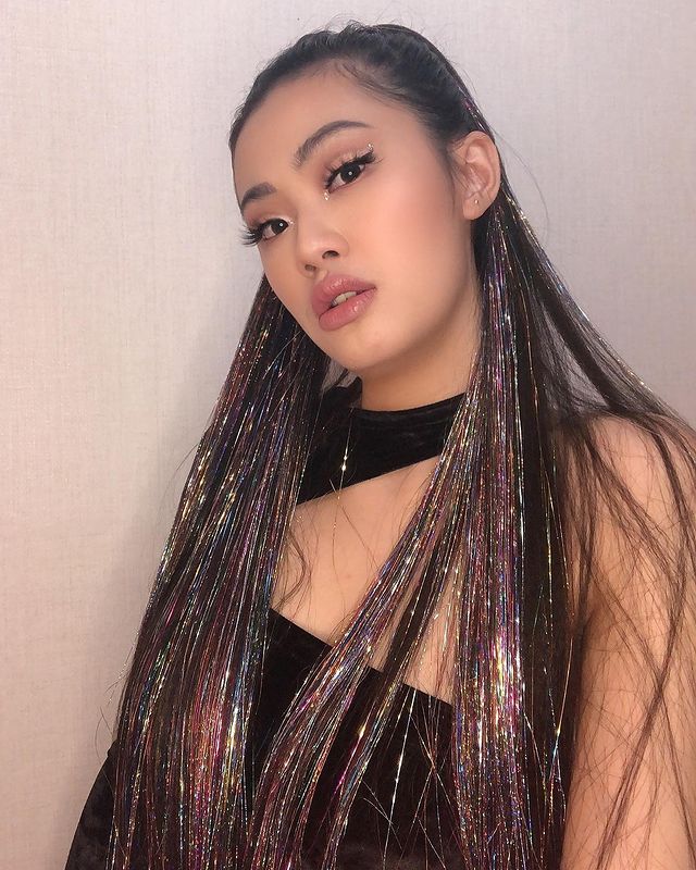 tinsel-hair Selfie de uma mulher. Ela usa um top cropped preto, maquiagem com cílios postiços e delineador preto e cabelo solto com fios de glitter. Ela olha para a câmera e não sorri.