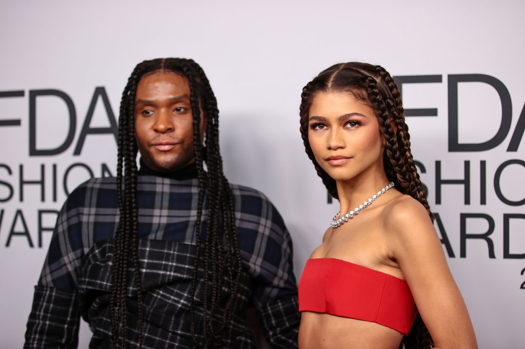 Foto da Zendaya e Law Roach no tapete vermelho. Law Roach está do lado esquerdo da foto, usa uma roupa quadriculada em tons de preto e cinza, cabelo com tranças, olha para o lado e não sorri. Zendaya está do lado direito da foto, usa top reto vermelho, colar prata, trança no cabelo, olha para o lado e não sorri.