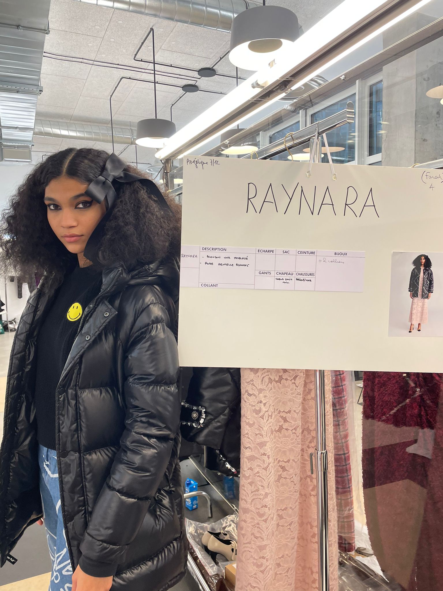 Raynara Negrine nos bastidores do desfile Metiers D'Art da Chanel. Ela posa à esquerda, ao lado de um painel com seu nome e o look que usa no desfile, de jaqueta puffer preta, camiseta preta, calça jeans e laço no cabelo.