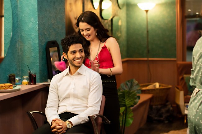 The Royal Treatment Laura Marano como Izzy e Mena Massoud como princípe Thomas em Tratamento de Realeza, da Netflix; ele está sentando sorrindo e olhando levemente para o lado enquanto ela está em pé atrás dele mexendo em seu cabelo para arrumá-lo