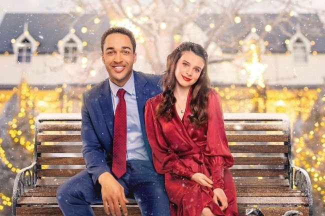 adotando-amor-no-natal Um homem de terno azul e gravata vermelha sentado um banco no parque cheio de neve ao lado de uma mulher com vestido e batom vermelho.