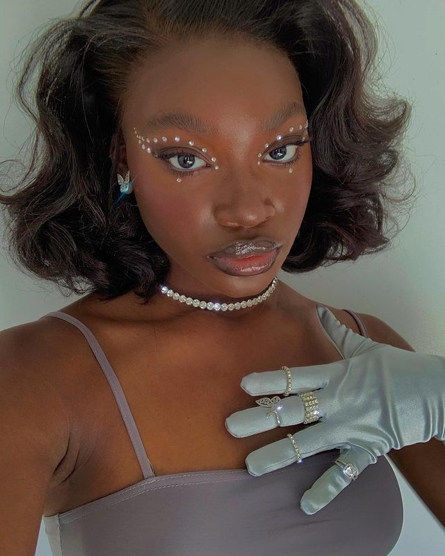 delineado-com-brilhos-aplicados Selfie de uma mulher. Ela usa um top cropped de alça, luvas com anéis dourados, colar de brilhos, brinco prata, cabelo solto e maquiagem com aplique de brilhos nos olhos. Ela olha para a câmera e não sorri.