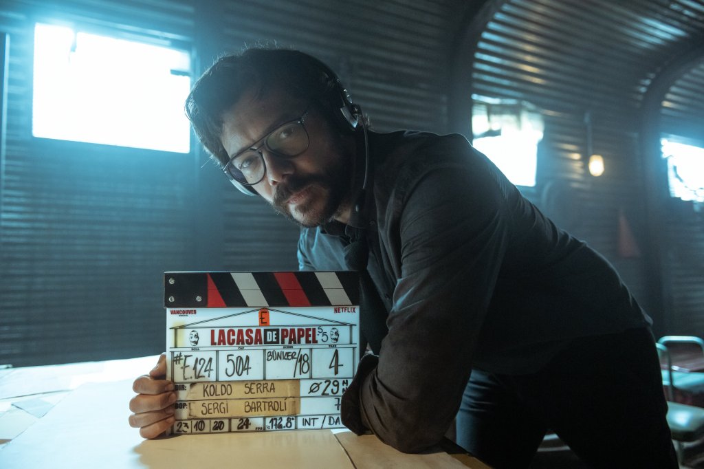 O Professor de La Casa de Papel nos bastidores do set da série segurando uma claquete enquanto se apoia em uma mesa; ele está com uma escuta no ouvido, camisa cinza, calça preta, óculos e tem expressão séria