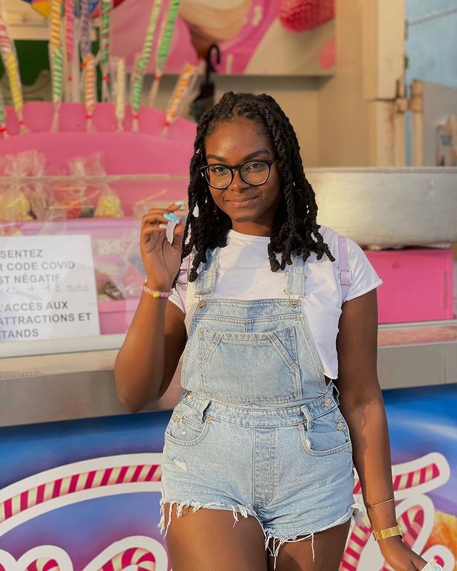 Foto de uma mulher em frente a uma banca de doces. Ela usa uma camiseta branca e jardineira curta jeans. Ela olha para a câmera e sorri.