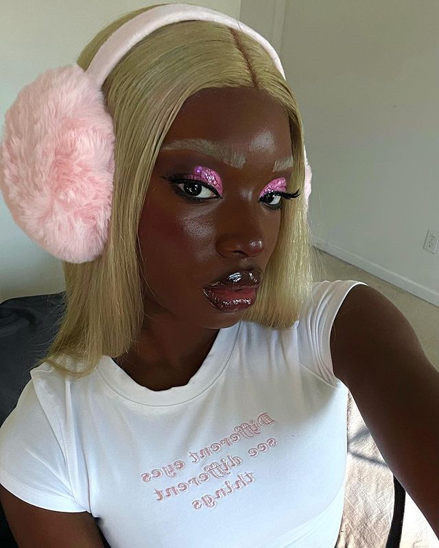 make-com-glitter-rosa Selfie de uma mulher. Ela usa uma baby tee branca, cabelo loiro liso, protetor de orelhas de pelúcia, pele natural, sobrancelha descolorida, maquiagem com glitter rosa e gloss. Ela olha para a câmera e não sorri.