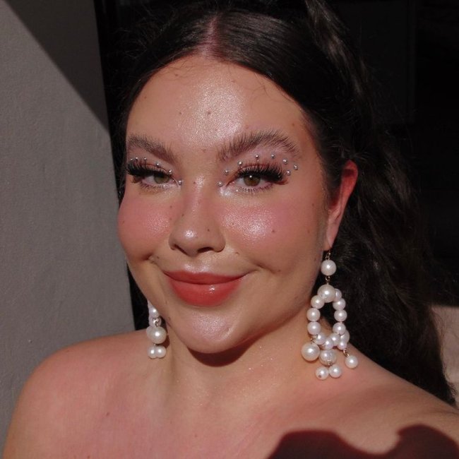 maquiagem-com-delineado-de-brilhos Selfie de uma mulher. Ela usa um brinco grande de perólas, cabelo preso em um rabo de cavalo, maquiagem básica iluminada e um delineado feito com aplicações de brilhos. Ela olha para a câmera e sorri levemente.