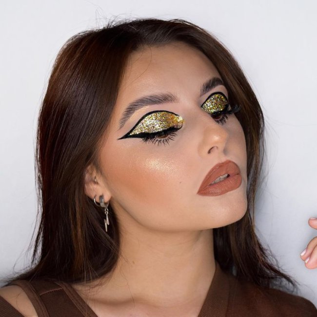 maquiagem-com-glitter-dourado Foto do rosto de uma mulher. Ela usa uma blusa marrom, cabelo solto, brinco de argola pequeno e maquiagem com pele glow, batom marrom e olho com glitter dourado na pálpebra e delineado preto. Ela olha para o lado e não sorri para a foto.