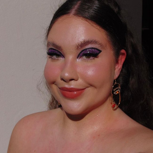 maquiagem-com-glitter-roxo Selfie de uma mulher. Ela usa o cabelo preso em um rabo de cavalo e maquiagem com a pele leve e olhos com glitter roxo e delineado preto.