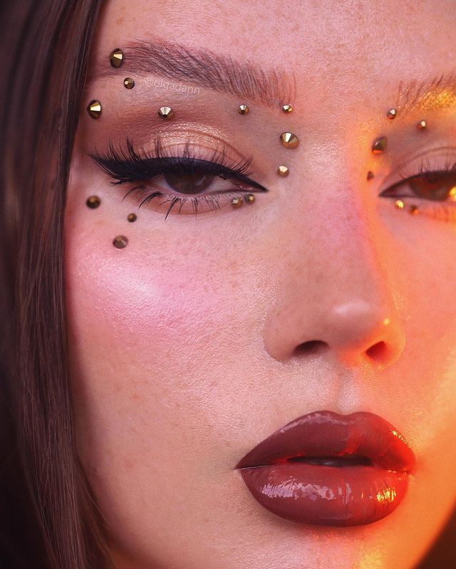 maquiagem-com-strass Foto close no rosto de uma mulher. Ela usa uma maquiagem básica com gloss, delineado preto e aplicações douradas. Ela olha para a câmera e não sorri.