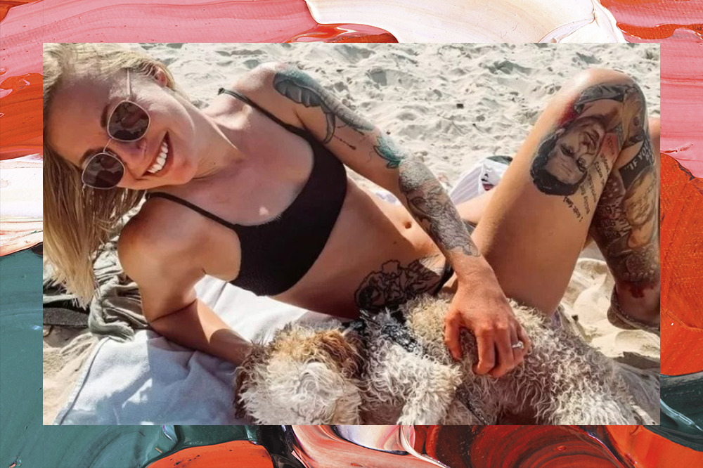 Foto de uma mulher loira, deitada na areia da praia com um cachorro bege, vestindo um biquíni preto e ostentando várias tatuagens