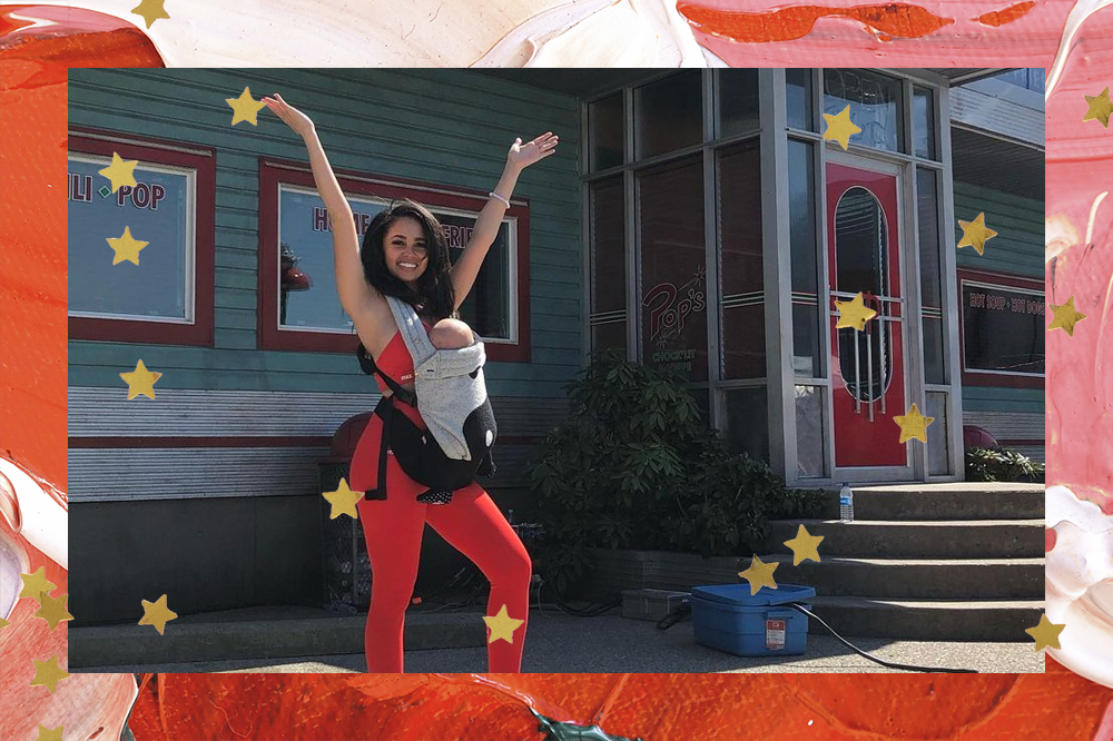 Vanessa Morgan com seu bebê no set de Riverdale; ela está com os braços levantados e sorrindo do lado de fora do restaurante Pop's; a margem é uma textura de tintas nas cores vermelho, rosa, branco, vermelho e verde com estrelas amarelas como decoração