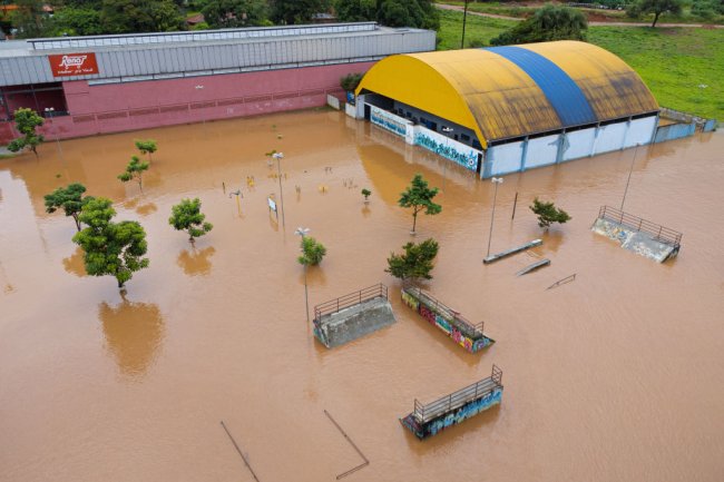 Chuvas na Bahia e em Minas Gerais: saiba como ajudar as vítimas Foto aérea de uma cidade mineira completamente alagada. Dá para ver o ginásio de uma escola com água até o teto praticamente