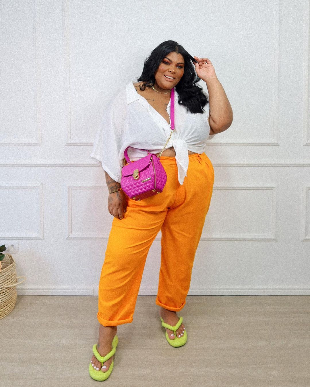 chinelo-acolchoado-relaxaaifofa Mulher sorrindo com uma das mãos no cabelo usando camisa branca, calça laranja, bolsa rosa e chinelo amarelo neon