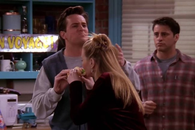 friends-joey Mulher loira comendo um sanduíche. Um homem segurando o lanche e outro olhando do lado.