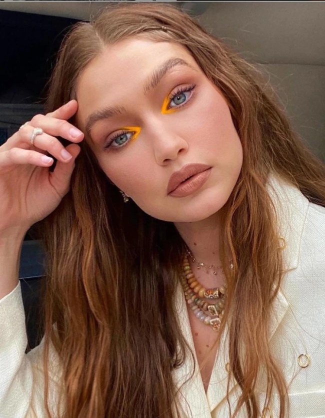 gigi-hadid-maquiagem-canto-interno-neon Foto da Gigi Hadid. Ela usa uma camisa branca, mix de colares, cabelo solto e maquiagem com batom nude e sombra laranja neon no canto interno dos olhos. Ela está com a mão direita na cabeça, olha para a câmera e não sorri.