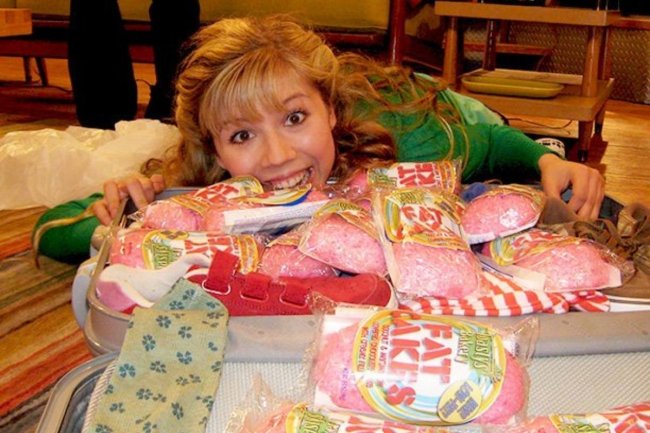 icarly-bolo-gordo Menina de blusa verde e cabelos loiros ao lado de uma mesa cheia de bolos rosas.