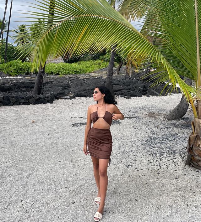 Foto de uma mulher na praia. Ela usa um top cropped marrom de amarração, saia marrom com detalhe franzido nas laterais, sandália branca e óculos de sol. Ela olha para o lado e não sorri.