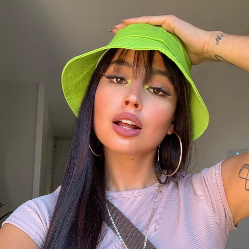 neon-maquiagem-canto-interno-irisloveunicorns Selfie de uma mulher. Ela usa um bucket hat verde, cabelo solt, brinco de argola, camiseta lilás e maquiagem com delineador e ponto de luz neon no canto interno dos olhos. Ela está com a mão esquerda na cabeça, olha para a câmera e não sorri.