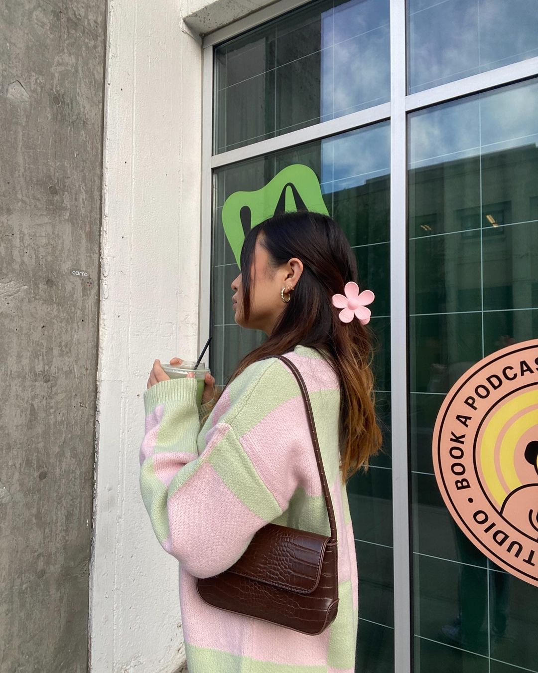 piranha-cabelo-flor-chiyobabe Garota de lado usando cardigan rosa e verde e bolsa marrom pendurada no ombro. Seu cabelo está preso com uma piranha em formato de flor rosa.