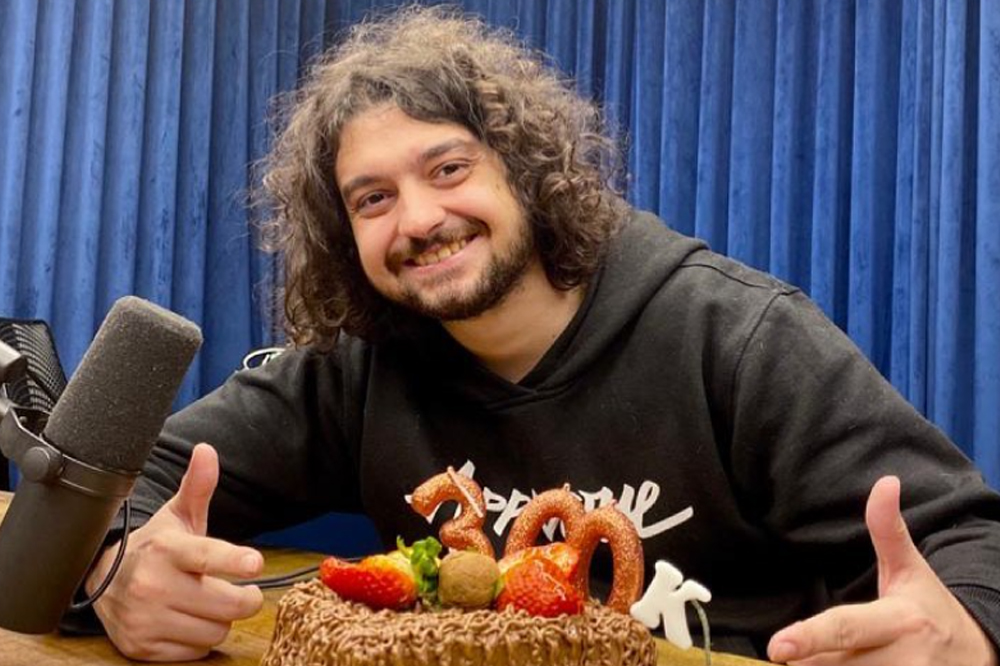 Foto do podcaster Monark. Ele sorri para a câmera, tem barba e cabelo comprido