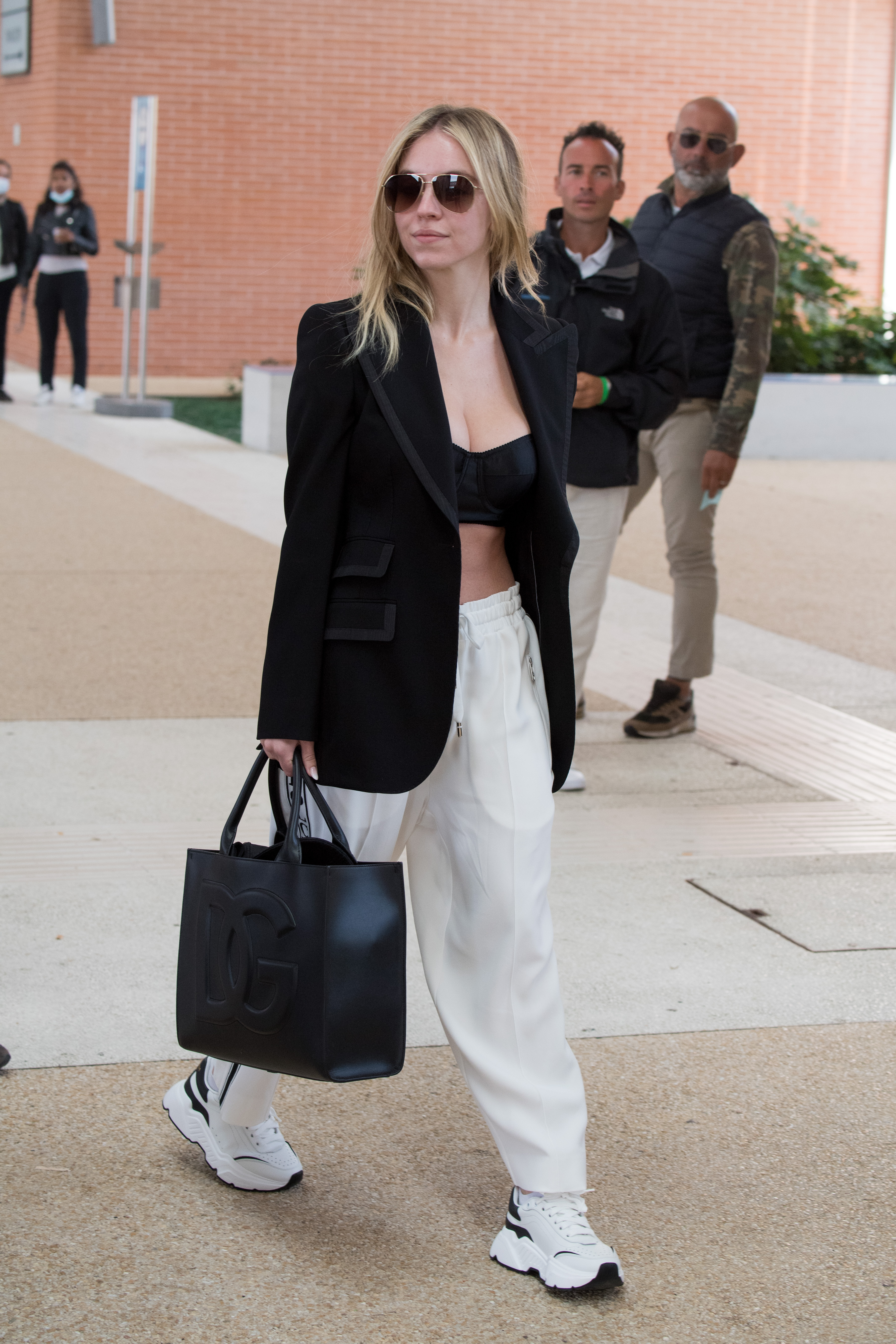 Celebrity Sightings – The 78th Venice International Film Festival Sydney Sweeney andando na rua e usando look com blazer e top cropped pretos, calça e tênis brancos e segurando uma maxibolsa preta, além de estar usando óculos de sol