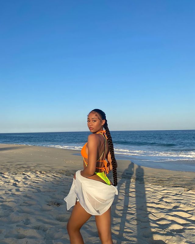 Foto de uma mulher na praia. Ela usa um biquíni laranja, segura uma camisa branca e está com um penteado de rabo de cavalo baixo trançado. Ela está de lado, olha para a câmera e não sorri para a foto.