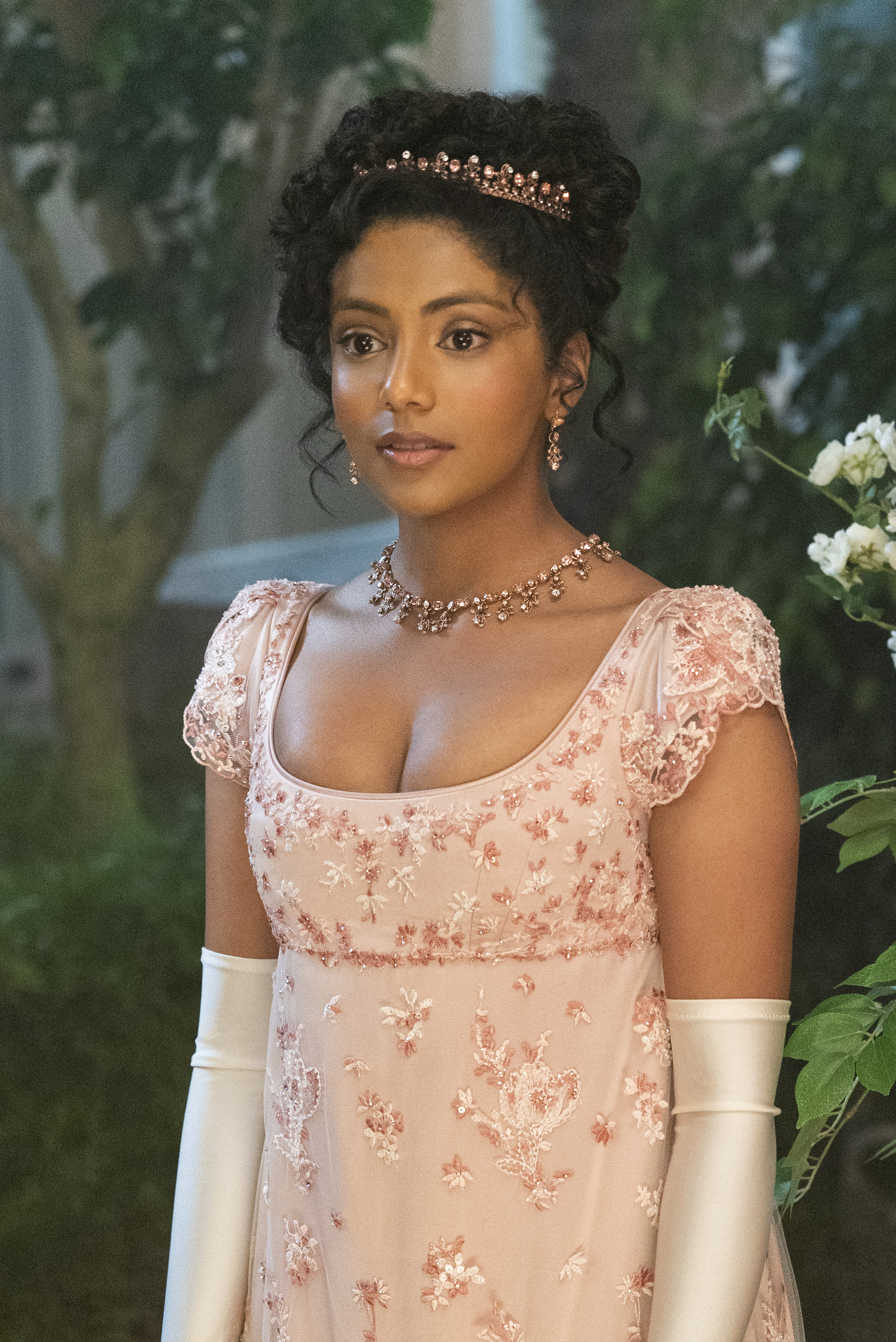BRIDGERTON_201_Unit_04483RC Imagem mostra Charithra Chandran caracterizada como Edwina Sharma usando joias de ouro rosa e vestido bordado com os mesmo elementos e luvas brancas em cena do 1º episódio da segunda temporada de Bridgerton.