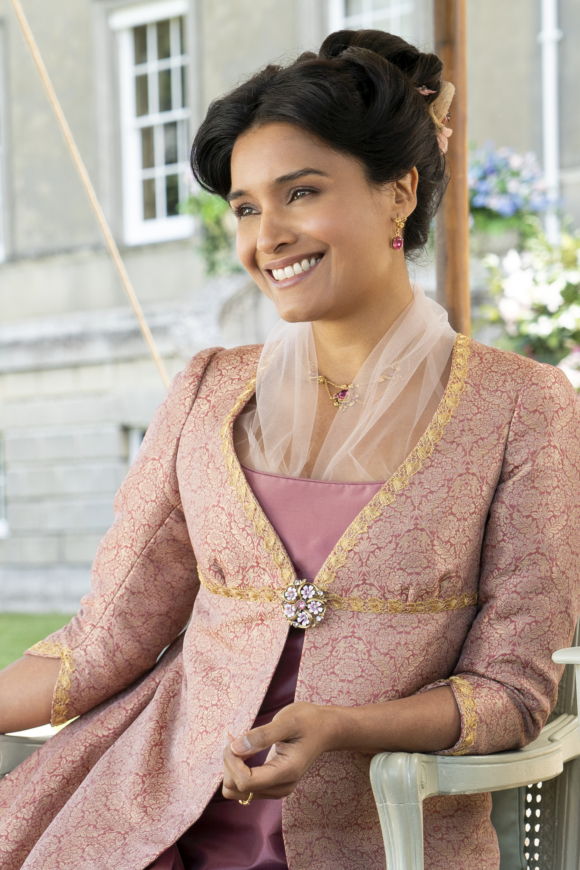 BRIDGERTON_203_Unit_02149RC Imagem mostra Shelley Conn caracterizada como Mary Sharma, sentada e sorrindo em cena da 2ª temporada de Bridgerton.