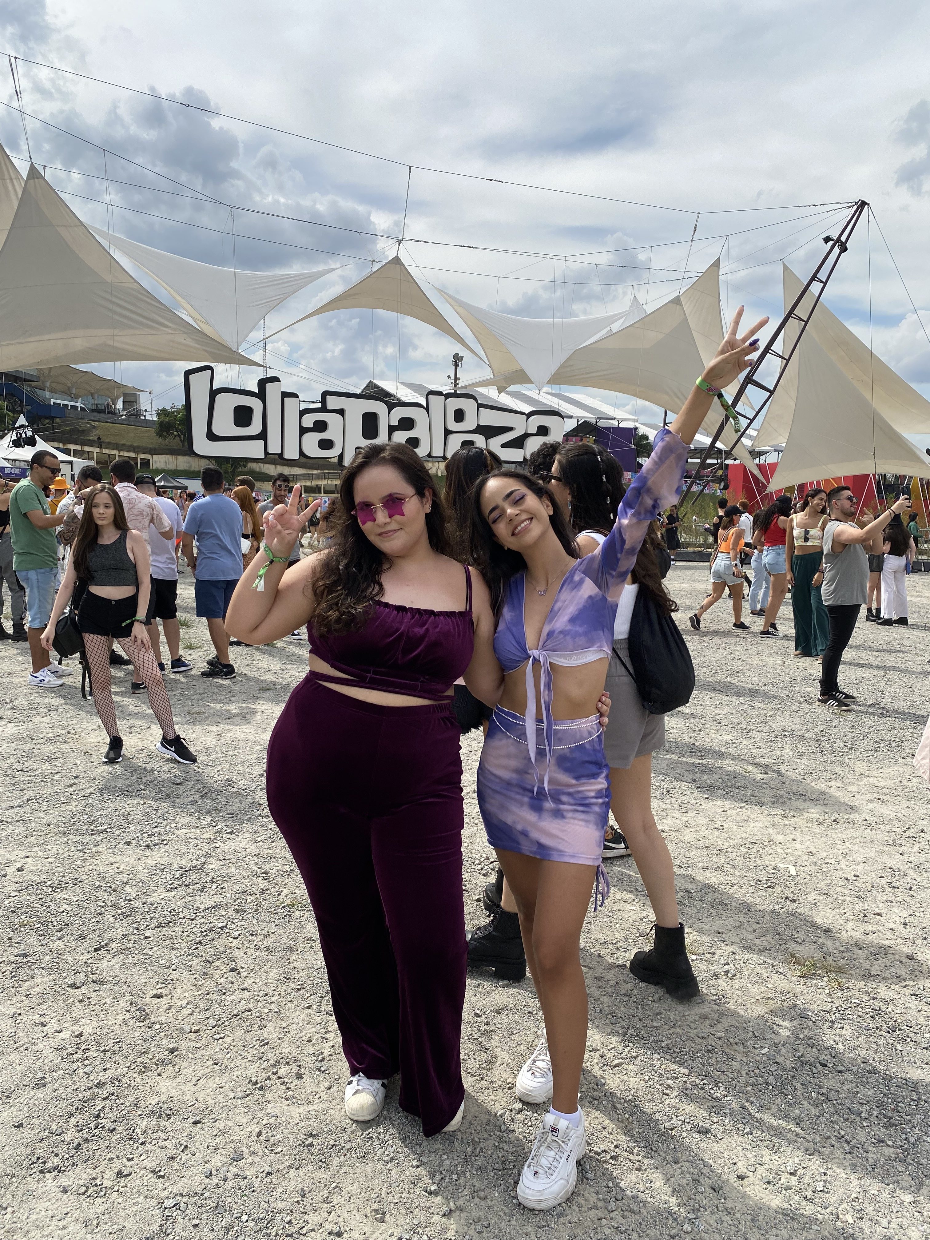 look-lollapalooza-conjuntinho Duas garotas em frente ao letreiro do Lollapalooza usando conjuntinho. À esquerda, menina de calça e top de veludo roxo. À direita, menina de top e saia tie-dye roxo.