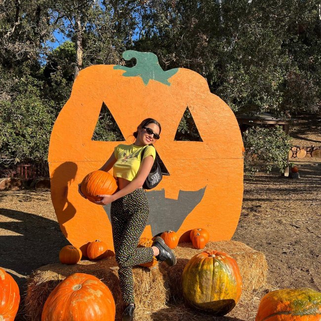olivia-rodrigo-baby-tee Foto da cantora Olivia Rodrigo em um local aberto com abóboras no chão. Ela usa uma baby tee amarela, calça estampada e calça um oxford. Ela segura uma abóbora, olha para a câmera e sorri.