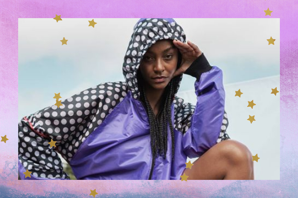 Montagem em fundo degradê roxo e azul com estrelinhas douradas. Garota usando corta-vento preto e branco de bolinhas com detalhe roxo na parte da frente, com uma das mãos apoiadas na testa e o braço no joelho.