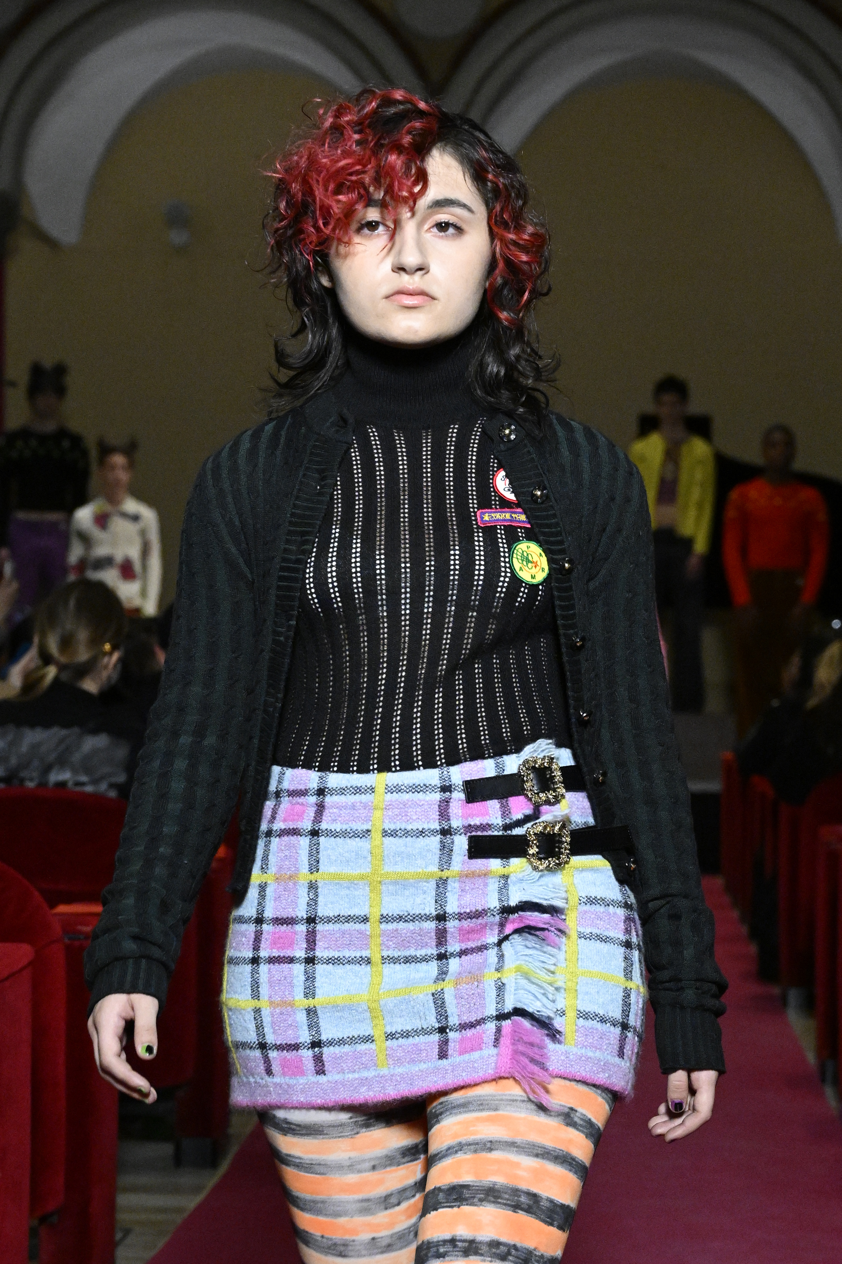 Modelo com cabelo preto e vermelho, tricô de manga comprida preto e saia xadrez em desfile da Cormio na semana de moda de Milão