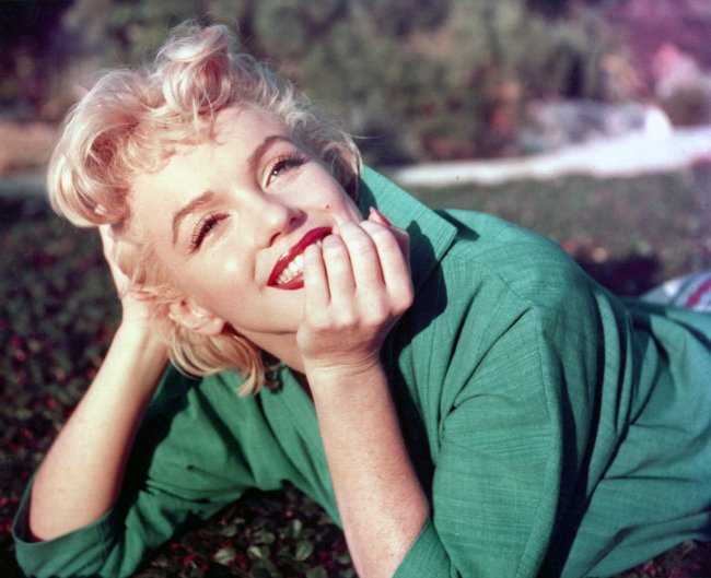tendencia-sobrancelha-anos-50 Foto da atriz Marilyn Monroe em 1954. Ela está deitada na grama da um parque, usa uma camisa verde, cabelo solto, maquiagem básica com batom vermelho. Ela olha para frente, com a mão no queixo e sorri.